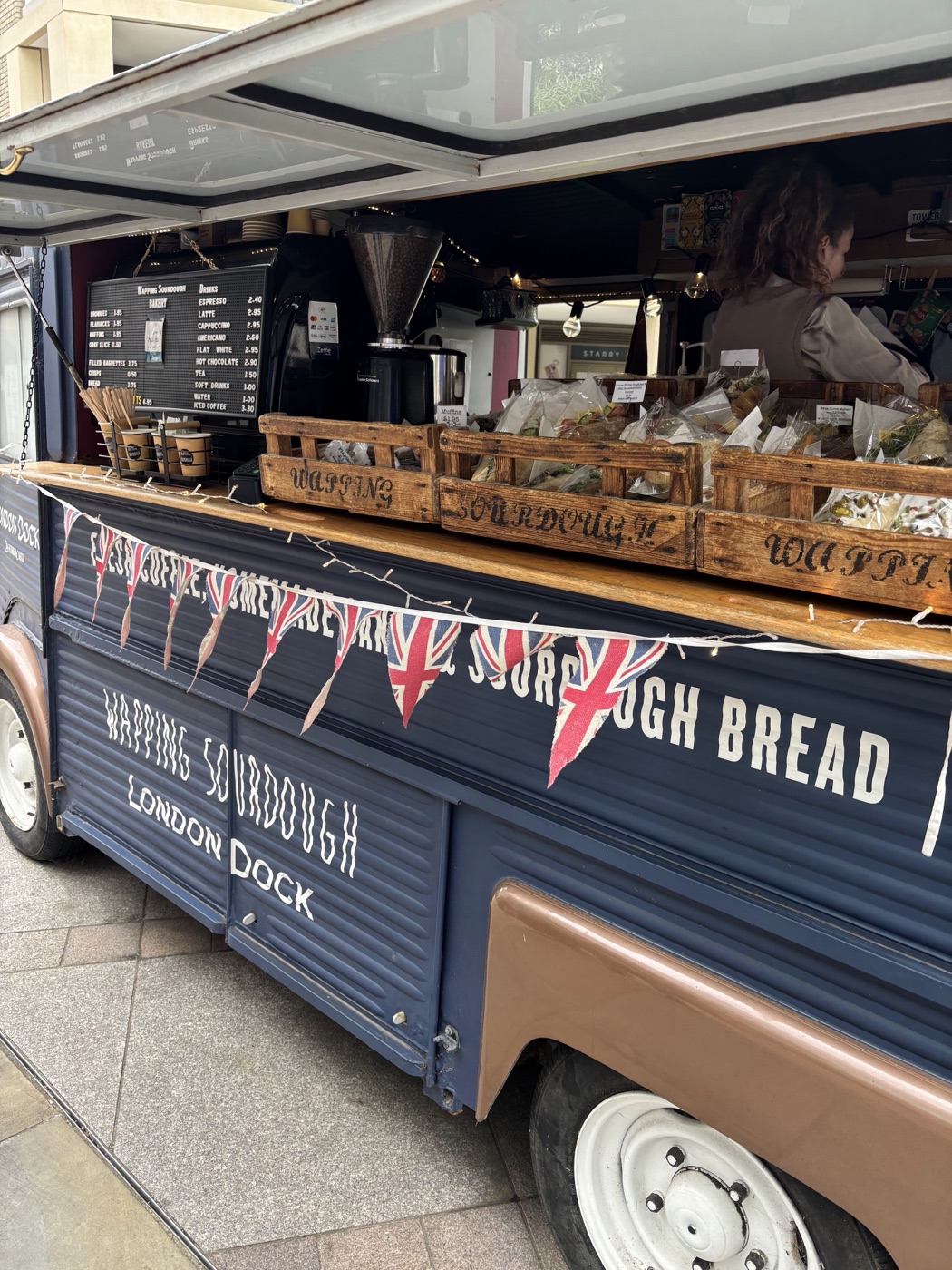 The vintage blue Wapping Sourdough van on Arrival Square, hand-painted lettering along the side, bread crates and bunting across the counter